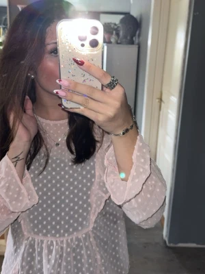 Ljusrosa prickig blus med volanger - Söt ljusrosa blus med vita prickar och volangdetaljer. Blusen har långärmad design och är tillverkad i ett tunt, transparent material som ger en romantisk och trendig look. Perfekt att styla med jeans eller kjol för en chic outfit.