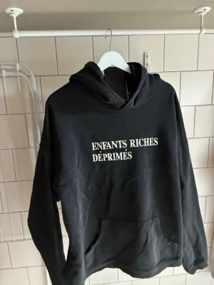 Enfants Riches Déprimés - Hoodie - Svart hoodie från Enfants Riches Déprimés med vit text på bröstet. Klassisk passform med stor huva och känguruficka framtill. Tillverkad i mjukt material som känns skönt mot huden. Perfekt för dig som gillar streetwear och statement-plagg. Ny med lappar