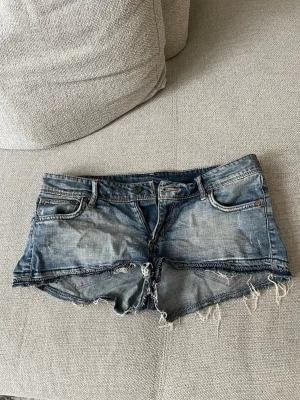 Blå jeansshorts med fransig kant - Säljer ett par blå denimshorts med rå, fransig kant och slitna detaljer. Klassisk femficksmodell med knapp och dragkedja framtill. Perfekta för sommaren och ger en avslappnad vibe.