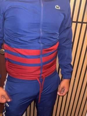 Lacoste tracksuit - Säljer en snygg blå träningsdress från Lacoste med röda och blå ränder över magen. Jackan har dragkedja och resår vid ärmslut, byxorna har dragsko i midjan. Perfekt för sport eller chill. Klassisk krokodil-logga på bröstet och byxan.