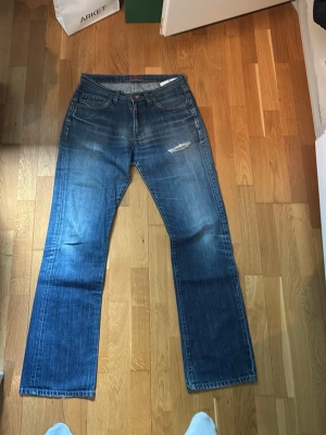 Blå raka jeans från G-Star - Säljer ett par klassiska blå jeans från G-Star med rak passform och fem fickor. Jeansen har slitningar på låret och snygg tvättad look. Tillverkade i denim med detaljerade sömmar och patch bak i midjan.