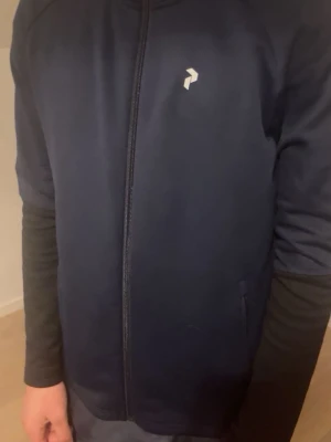 Mörkblå hoodie från Peak Performance - Snygg mörkblå hoodie från Peak Performance med dragkedja framtill och diskret logga på bröstet. Huva med textdetalj runt kanten. Tillverkad i mjukt material som känns skönt mot huden. Perfekt för en sportig och avslappnad look.