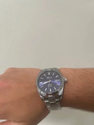 Blå och silverfärgad Domossi klocka+ plus armbandsverktyg  - Stilren klocka från Domossi med blå urtavla och datumvisning. Armbandet är i silverfärgad metall och har en klassisk länkdesign. Klockan har tydliga markeringar och kvartsverk. Perfekt accessoar för dig som gillar en clean look.