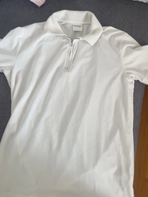 Vit Piké  - Klassisk vit piké från Selected Homme med kort ärm och krage. Pikén har en snygg dragkedja framtill som detalj och är gjord i mjuk bomull. Perfekt till jeans eller kjol för en clean look. Den har dock en fläck på baksidan av Pikén. 