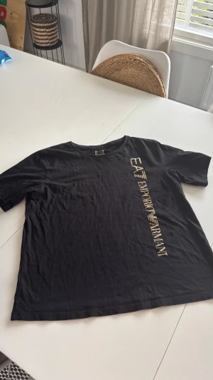 Svart EA7 Emporio Armani t-shirt - Svart t-shirt från EA7 Emporio Armani med guldig logga tryckt vertikalt på framsidan. Klassisk rund halsringning och korta ärmar. Tillverkad i mjuk bomull för en bekväm känsla och stilren look.