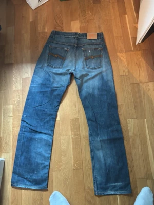 Blå raka jeans från Nudie Jeans - Säljer ett par blå raka jeans från Nudie Jeans (modell: straight sven) med slitningar och hål på benen för en cool, avslappnad look. Jeansen har klassiska fem fickor och kontrastsömmar på bakfickorna. Perfekta för dig som gillar streetwear och vintage vibe.