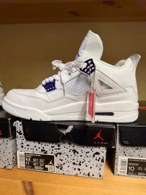 Air Jordan 4 Retro Vit/Lila - Säljer ett par Air Jordan 4 Retro sneakers i vitt läder med lila detaljer och meshpaneler. Klassisk Jumpman-logga på hälen och tungan. Skorna har högt skaft och synlig Air-enhet i sulan. Perfekta för dig som vill sticka ut med stilrena och ikoniska sneakers.
