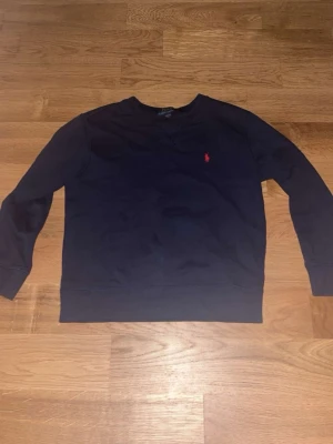 Mörkblå crewneck från Ralph Lauren - Mörkblå sweatshirt från Ralph Lauren med klassisk rund halsringning och röd broderad logga på bröstet. Tröjan har långa ärmar och ribbade muddar vid ärmslut och nederkant. Perfekt för en avslappnad och stilren look.