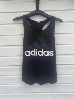 Svart linnetopp från Adidas - Snygg svart linnetopp från Adidas med stort vitt logotryck framtill. Modellen är ärmlös med brottarrygg och har en sportig vibe, perfekt för träning eller chill. Tillverkad i mjukt material som känns skönt mot huden. Väl använd storlek vet ej