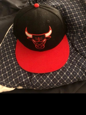 Chicago Bulls keps Mitchell & Ness - Snygg snapback-keps från Mitchell & Ness med Chicago Bulls-logga framtill. Kepsen är svart med röd skärm och röd knapp på toppen. Justerbar baktill med röd plastrem. Undersidan av skärmen har ett tryck med Bulls-tema. Perfekt för dig som gillar streetwear och sport.