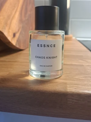 ESSNCE Chaos Knight Eau de Parfum - En stilren parfymflaska från ESSNCE med doften Chaos Knight. Flaskan är cylindrisk i genomskinligt glas med svart lock och vit etikett med svart text. Modern och minimalistisk design som passar perfekt på parfymhyllan.