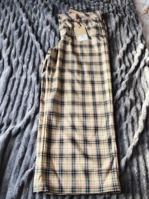 Rutiga byxor i beige och blått - Snygga byxor med rutigt mönster i beige, blått och grönt. Byxorna har raka ben och är tillverkade i ett mjukt tyg som känns skönt mot huden. Perfekta för dig som vill sticka ut med ett klassiskt men trendigt mönster.