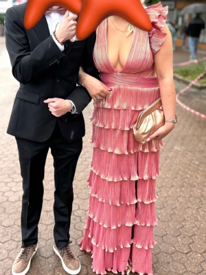 Rosa långklänning med volanger - Säljer en rosa långklänning med djup v-ringning, volangdetaljer och snörning i ryggen. Klänningen har plisserade lager och axelvolanger som ger en unik look. Perfekt för bal eller festliga tillfällen. Materialet känns som polyester. Använd 1 gång på balen iår 