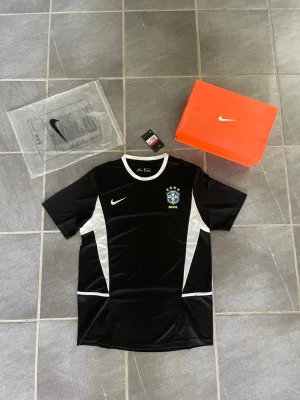 Svart Brasilien Nike fotbollströja - Snygg svart fotbollströja från Nike med Brasilien-emblem på bröstet och vita detaljer längs sidorna och kragen. Tillverkad i lätt och ventilerande material, perfekt för träning eller match. Klassisk passform och korta ärmar.