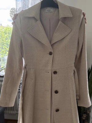 Beige kappa från ONLY med bälte - Stilren beige kappa från ONLY med klassisk krage, knappar framtill och tillhörande bälte i midjan. Kappan har två fickor och är tillverkad i ett vävt tyg med diskret struktur. Perfekt för dig som gillar en tidlös och elegant look.