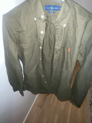 Olivgrön skjorta från Ralph Lauren - Snygg olivgrön skjorta från Ralph Lauren med klassisk krage och knappar framtill. Skjortan har lång ärm och den ikoniska loggan broderad i orange på bröstet. Tillverkad i bomull för en skön känsla.