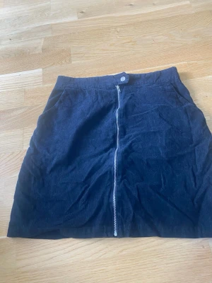 Svart manchesterkjol från SHEIN - Säljer en svart kortkjol i manchester från SHEIN. Kjolen har en rak passform, praktiska fickor och en synlig dragkedja framtill som detalj. Perfekt för dig som gillar en enkel men trendig stil.
