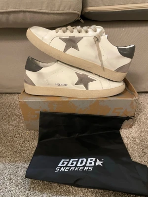 GGDB Superstar vita sneakers - Snygga vita GGDB Superstar sneakers med grå stjärna på sidan och grå häl. Skorna har beige sula och vita snören. Tillverkade i läder med detaljer i mocka och klassisk låg siluett. Perfekta för dig som gillar streetstyle och vill ha något unikt.