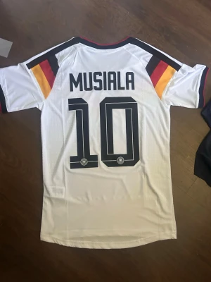 Tysklands hemma matchtröja Musiala 10  - Player-version repelica. Snygg vit Tyskland matchtröja från Adidas med Musiala och nummer 10 på ryggen. Tröjan har klassiska svarta, röda och gula detaljer över bröstet och axlarna, samt officiellt landslagsmärke och Adidas-logga framtill. Tillverkad i lätt och ventilerande material.