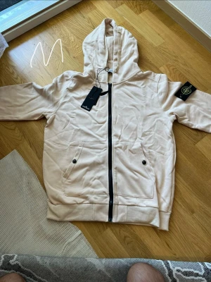 Beige hoodie från Stone Island - Snygg beige hoodie från Stone Island med dragkedja framtill och klassisk logopatch på vänster ärm. Hoodien har två fickor med knappstängning och en stor huva. Perfekt för en avslappnad streetwear-look.