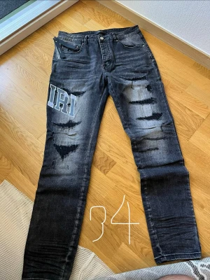 Svarta distressed jeans med tryck - Säljer ett par svarta jeans med slitningar och tryck i vitt och blått på både fram- och baksida. Jeansen har raka ben, fem fickor och coola detaljer som ger en streetwear-vibe. Perfekta för dig som gillar en edgy stil.
