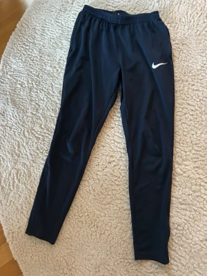 Mörkblåa träningsbyxor från Nike - Snygga mörkblåaträningsbyxor från Nike med vit logga på vänster lår. Byxorna har smal passform, resår i midjan och är tillverkade i ett lätt och stretchigt material som passar perfekt för sport och träning. Byxorna har inga hål eller så. De passar tyvärr inte mig längre. Storlek S