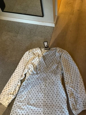 Vit prickig omlottklänning - Vit klänning med svarta prickar och omlott-design. Djup v-ringning och långa ärmar med lätt puff vid axlarna. Klänningen har en feminin siluett och är perfekt för dig som gillar en klassisk men trendig look.