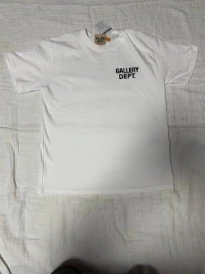 Vit t-shirt från Gallery Dept. - Vit t-shirt från Gallery Dept. med svart tryck på bröstet och stor text på ryggen. Klassisk passform och rund halsringning. Perfekt för en clean och stilren look.