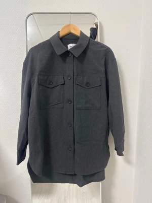 Svart overshirt från Zara - Stilren svart overshirt från Zara med klassisk krage och raka linjer. Två bröstfickor med lock och knappar framtill ger en cool utility-vibe. Perfekt att bära öppen eller stängd över en t-shirt. Materialet känns mjukt och har en matt finish.