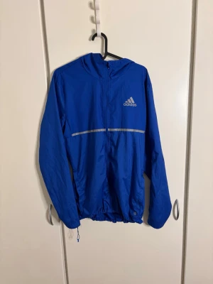 Blå vindjacka från Adidas - Snygg blå vindjacka från Adidas med huva och reflekterande detalj över bröstet. Jackan är lätt, har dragkedja framtill och justerbar nederkant. Perfekt för löpning eller när du vill ha en sportig look.