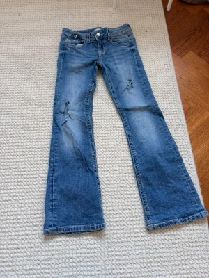 Blå bootcut jeans med slitningar - Säljer ett par blå bootcut jeans med slitningar på benen och klassisk femficksdesign. Jeansen har ljus tvätt och råa detaljer som ger en cool look. Perfekt för dig som gillar en avslappnad och trendig stil. Fråga om mer bilder. Storlek 152. 