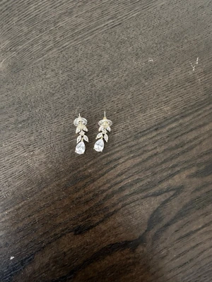 Senso di Donna  - Snygga örhängen med droppformade kristaller och små blad i guldton. De har en elegant design med gnistrande detaljer som verkligen sticker ut. Perfekta för dig som gillar att addera lite extra bling till din stil.