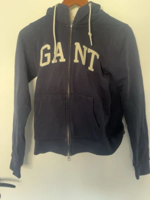 Mörkblå hoodie med dragkedja från GANT - Snygg mörkblå hoodie från GANT med vit broderad logga på bröstet. Modellen har dragkedja framtill, huva med vita snören och två fickor. Perfekt för en avslappnad och sportig stil.