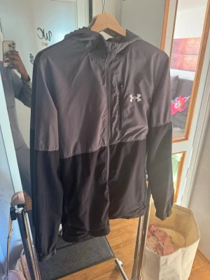 Svart vindjacka från Under Armour - Snygg svart vindjacka från Under Armour med huva och dragkedja framtill. Jackan har en bröstficka med dragkedja och logga på vänster sida. Tillverkad i lätt och vindtätt material, perfekt för blåsiga dagar. Enkel och stilren design som passar till det mesta.