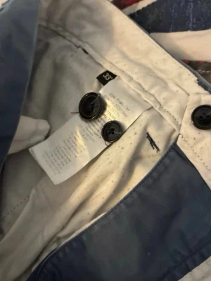 Blå chinos från Ralph Lauren - Snygga blå chinos från Ralph Lauren med klassisk passform och bälteshällor. Byxorna har två knappar i midjan och är tillverkade i bomull för en bekväm känsla. Perfekta för en stilren och avslappnad look.