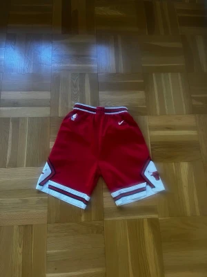 Röda Chicago Bulls basketshorts Nike - Snygga röda basketshorts från Nike med Chicago Bulls-logga på benen och vita detaljer. Shortsen har elastisk midja och sportig design, perfekta för dig som gillar basket eller streetwear.