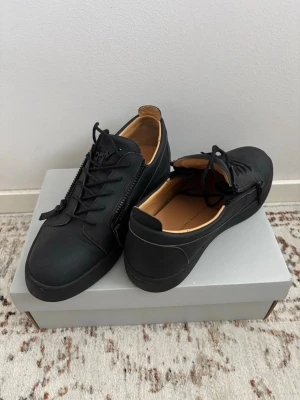Svarta sneakers från Giuseppe Zanotti - Stilrena svarta sneakers från Giuseppe Zanotti med mattsvart finish, svarta snören och dragkedjor på sidorna. Insidan är ljusbrun i läder och skon har en låg profil. Logan på vänstra skon har gått sönder.