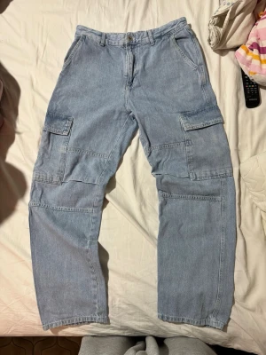 Ljusblå cargobyxor i denim - Säljer ett par ljusblå cargobyxor i denim med flera stora fickor på sidorna och klassisk rak passform. Byxorna har knapp och dragkedja framtill samt bälteshällor. Perfekta för en avslappnad och trendig look.