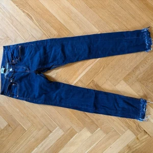 Mörkblå jeans från Lager  - Säljer ett par mörkblå jeans med smal passform och råa, fransiga benslut. Klassisk femficksmodell och knappgylf. Perfekta för en avslappnad och trendig look.