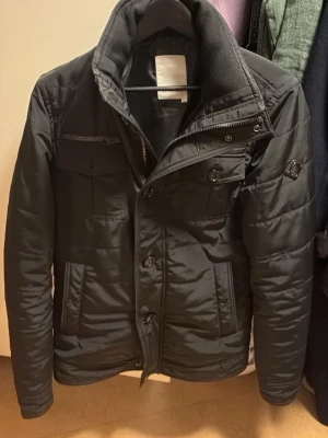 Svart field jacket från J.Lindeberg - Svart field jacket från J.Lindeberg med hög krage, flera fickor med både dragkedja och knappar samt snyggt emblem på ärmen. Jackan har en stilren look och är perfekt för vintern och våren och passformen är Xs.