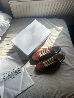 Maison Margiela bruna/burgundy - Maison Margiela sneakers i brunt läder och mocka med vita snören och beige innersida. Klassisk design med låg profil och detaljerad text på plösen. Perfekt för dig som vill ha något stilrent och exklusivt. Lite roligare färg som inte är så basic. Endast testade finns dustbag och box kvar! Hör av er vid eventuella frågor! Storlek 45 kan tänka mig att diskutera priset vid en snabb affär