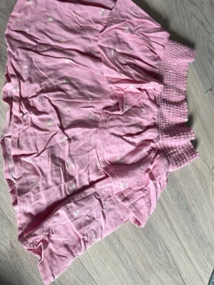 Rosa kortkjol med volang och små blommor - Söt rosa kortkjol med volangkant och små blommor. Kjolen har bred, smockad midja som ger en bekväm passform. Perfekt för vår och sommar när du vill ha en lätt och luftig stil.