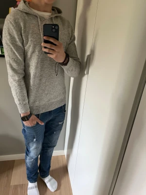 Ljusgrå stickad hoodie med huva - Säljer en stilren ljusgrå hoodie med huva och dragsnören. Tröjan är i mjukt stickat material och har ribbade muddar vid ärmslut och nederkant. Perfekt för en avslappnad och snygg look. Den har tyvärr ett litet håll på armen. Men det är igen sytt. Så syns knappt 