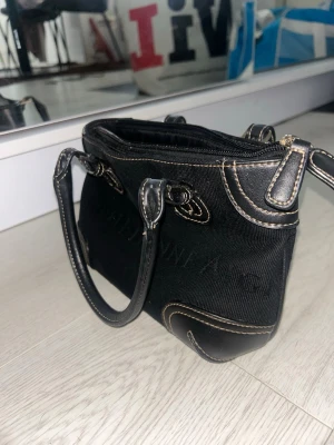 Svart handväska från Calvin Klein - Snygg svart handväska från Etienne Aigner i canvas med detaljer i läder och kontrastsömmar. Väskan har två handtag och dragkedja upptill. Perfekt storlek för det viktigaste och stilren design med diskret logga.