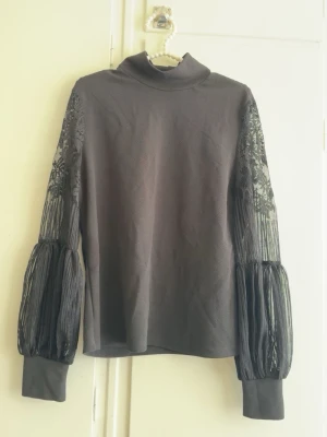 Grå polotopp med spetsärmar - Snygg grå polotopp med transparenta, långärmade spetsärmar och breda muddar. Ärmarna har en unik puffig design med plisserade detaljer som ger toppen en cool och trendig look. Perfekt för dig som vill sticka ut lite extra.