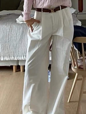 Vita vida kostymbyxor med pressveck - Säljer ett par vita kostymbyxor med hög midja och vida ben. Byxorna har pressveck framtill och bälteshällor i midjan. Perfekta för en clean och stilren look. Materialet ser ut att vara ett mjukt tyg med lite struktur.