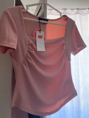 Rosa ribbad topp med fyrkantig ringning - Trendig ljusrosa ribbad topp med korta ärmar och snygg fyrkantig ringning. Toppen har rynkade detaljer framtill som ger en cool och modern look. Perfekt att styla med jeans eller kjol för en fräsch outfit.