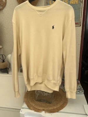 Gräddvit tröja från Polo Ralph Lauren - Klassisk beige tröja från Polo Ralph Lauren i 100% bomull. Tröjan har rund halsringning, ribbade muddar och den ikoniska lilla broderade loggan på bröstet. Perfekt för en clean och stilren look. Lite äldre stil på denna tröja, köptes på en Secondhand butik för 550kr då den är ganska unik pga av att den inte görs längre. Priset kan diskuteras!