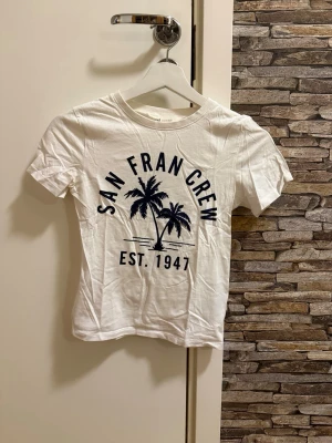 Vit t-shirt San Fran Crew - Vit t-shirt med tryck 'San Fran Crew Est. 1947' och palmmotiv i mörkblått. Klassisk rund hals och korta ärmar. Perfekt för en avslappnad stil och enkel att matcha med jeans eller shorts.