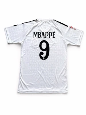 Real Madrid 24/25 Home Jersey – Mbappé #9 – Size XS - Up for sale is the iconic Real Madrid home shirt for the 24/25 season, featuring world superstar Kylian Mbappé and his number 9 on the back. This jersey features the classic white design with the subtle houndstooth-style pattern in the fabric. • Player: Kylian Mbappé (#9) • Size: XS (fits small adults or older juniors) • Condition: Excellent condition, very lightly used. • Details: Includes the La Liga sleeve patch and HP sponsor logo. 